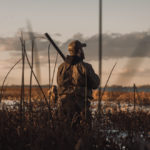 A duck hunter in camo stands in a marsh in the early morning